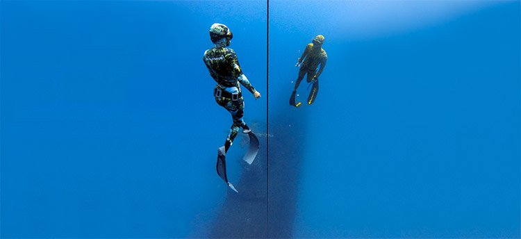 Female freediver wearing a blue and black wetsuit looking up to the surface of the water