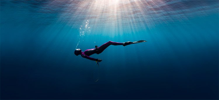 A male freediver in a black wetsuit holding a yellow snorkel ascending from a shipwreck in crystal clear water