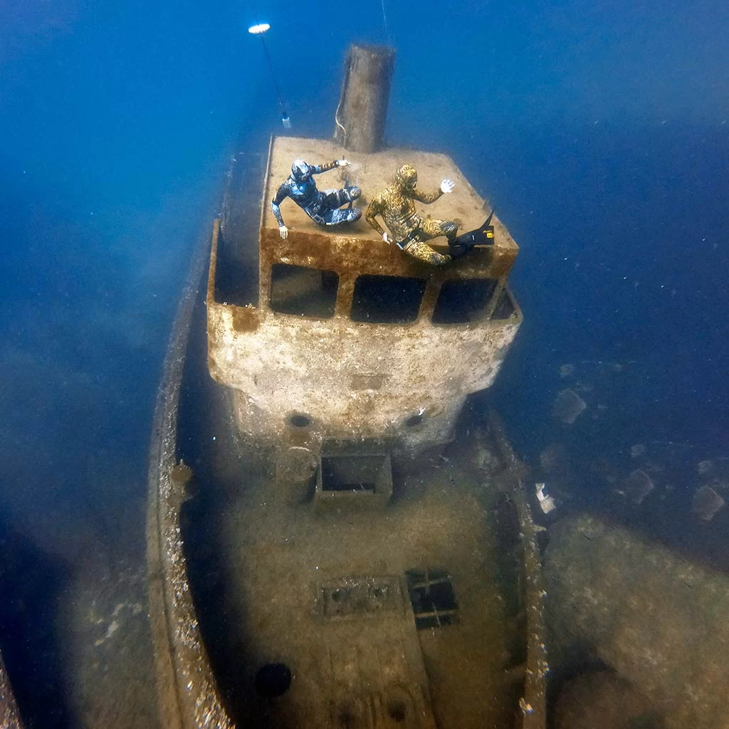 a couple of freedivers with camouflage wetsuits dancing on the top of a wreck