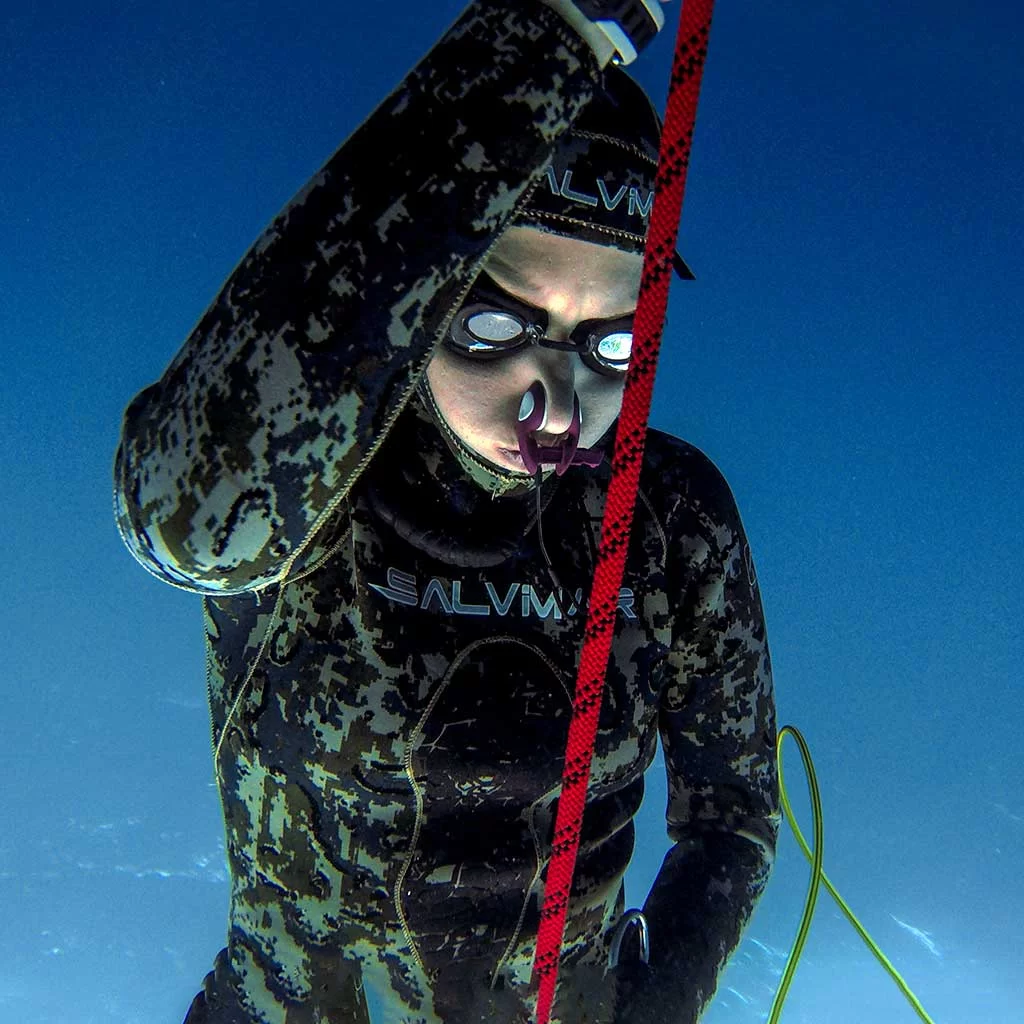 A freediver in a green wetsuit with fluid googles and a nose clip descending on the line